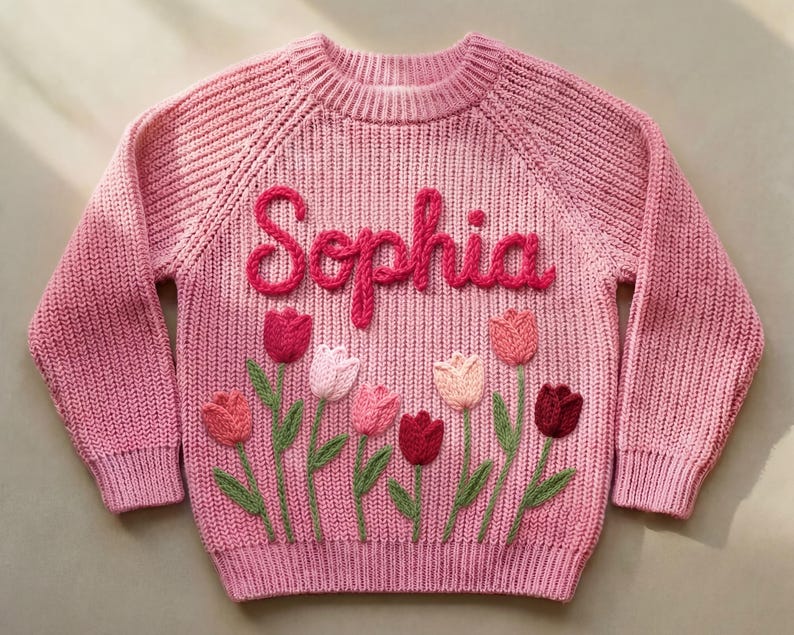 A pink knit sweater with the name "Sophia" embroidered in a cursive font. The sweater features embroidered tulips in shades of pink and red with green stems and leaves.