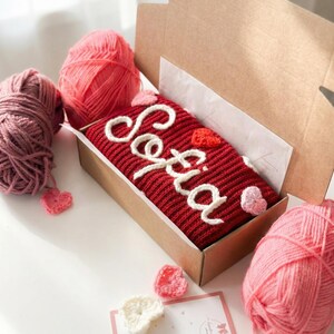 Puede incluir: Un art&iacute;culo tejido de color rojo con el nombre "Sofia" en escritura blanca, presentado en una caja de cart&oacute;n. El art&iacute;culo est&aacute; decorado con peque&ntilde;os corazones de ganchillo blancos y rosas. Tambi&eacute;n se ven ovillos de lana rosa y malva y una peque&ntilde;a tarjeta.