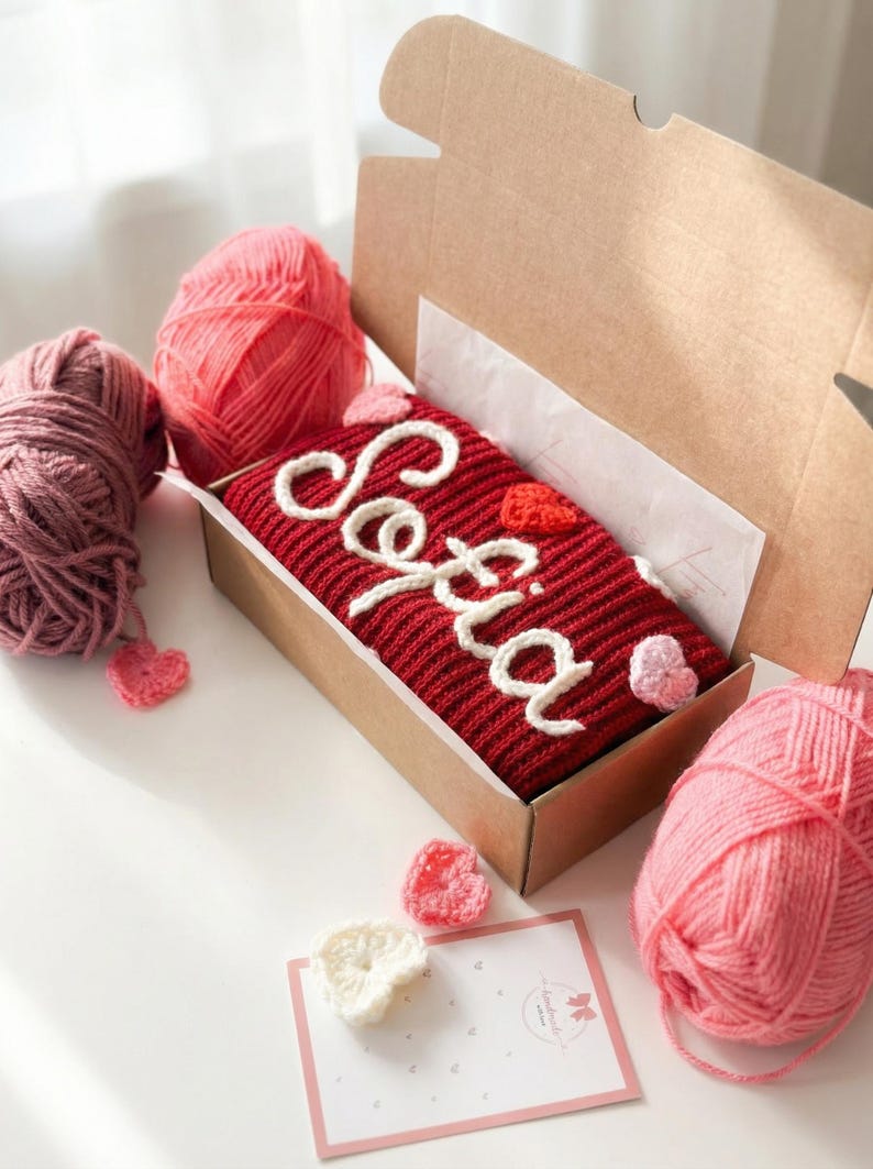 Puede incluir: Un art&iacute;culo tejido de color rojo con el nombre "Sofia" en escritura blanca, presentado en una caja de cart&oacute;n. El art&iacute;culo est&aacute; decorado con peque&ntilde;os corazones de ganchillo blancos y rosas. Tambi&eacute;n se ven ovillos de lana rosa y malva y una peque&ntilde;a tarjeta.