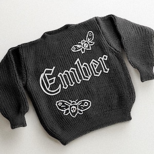 May include: Black knit sweater with the word "Ember" in white gothic lettering. Two white moth designs are embroidered on the sweater. The sweater has long sleeves and a ribbed hem and cuffs.