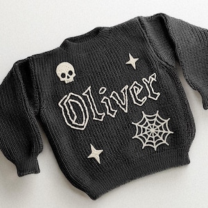 May include: Black knit sweater with the name "Oliver" in white, gothic-style lettering. The sweater features a skull, stars, and a spiderweb design. The sweater has long sleeves and a ribbed hem.