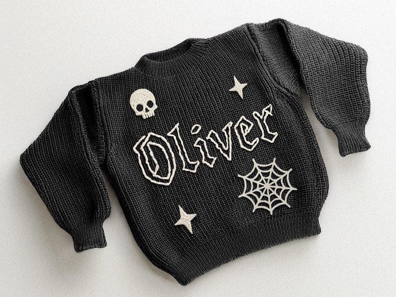 May include: Black knit sweater with the name "Oliver" in white, gothic-style lettering. The sweater features a skull, stars, and a spiderweb design. The sweater has long sleeves and a ribbed hem.