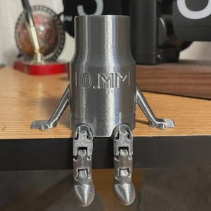May include: A gray 3D-printed figurine resembling a 10mm bullet casing with articulated legs and arms. The text "10.MM" is embossed on the casing. The figurine is sitting on a wooden surface.