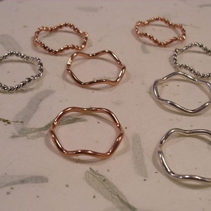 May include: A collection of eight silver and copper rings with a wavy or twisted design. The rings are arranged on a white surface.
