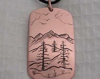 Puede incluir: Un collar con un colgante de cobre con un diseño grabado a mano de un paisaje de montaña con tres pinos. El colgante está unido a un cordón negro.