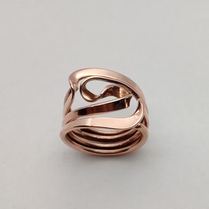 Puede incluir: Un anillo de oro rosa con un diseño único y abstracto. El anillo está hecho de múltiples bandas que están entrelazadas y tienen un acabado liso y pulido.