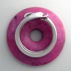 May include: A pink donut-shaped stone with a silver ring wrapped around it. The ring is open and has a smooth, rounded edge.
