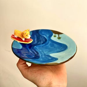 May include: A ceramic dish featuring a blue and white ocean scene with a small, yellow and orange dog figurine riding a red surfboard. The dish is held in a hand.