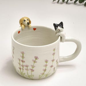 May include: White ceramic mug with a dog and cat design. The mug features a light pink floral pattern and red heart accents. A golden retriever figurine is perched on the rim, opposite a black and white cat figurine on the handle.