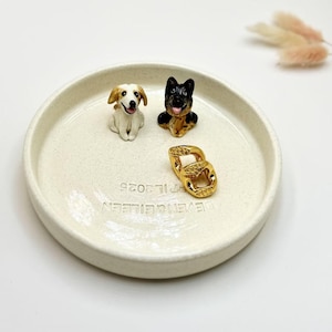 May include: A cream-colored ceramic dish with two small dog figurines and a gold-colored chain link. The dogs are a white and brown puppy and a black and tan German Shepherd. The dish has the text "DEVEN & EILEEN PT IL 2025".