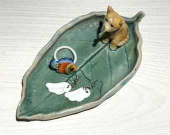 May include: A teal ceramic leaf-shaped trinket dish with a small brown cat figurine and two pairs of earrings. One pair features white cloud-shaped charms, and the other has a colorful fish design. The dish is on a white surface.