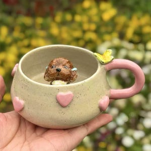 May include: A beige ceramic mug with a pink handle and pink hearts around the rim. A small brown dog figurine is inside the mug. A yellow butterfly is on the side of the mug.