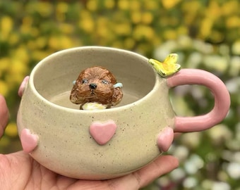 Puede incluir: Una taza de cerámica beige con asa rosa y corazones rosas alrededor del borde. Una pequeña figurita de perro marrón está dentro de la taza. Una mariposa amarilla está en el lado de la taza.