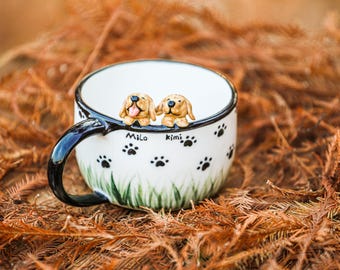 Może przedstawiać: Biały kubek ceramiczny z czarnym uchwytem i wzorem odcisków łap. Dwie małe ceramiczne figurki psów są umieszczone na krawędzi kubka. Figurki psów są brązowe i mają języki wystające. Kubek jest ozdobiony napisami "Milo" i "Kimi".