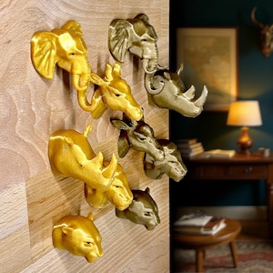 Può includere: Decorazioni da parete a forma di teste di animali, tra cui elefante, giraffa, rinoceronte e cervo, in tonalità oro e bronzo. I pezzi decorativi sono montati su una superficie in legno, aggiungendo un tocco di eleganza a tema safari.