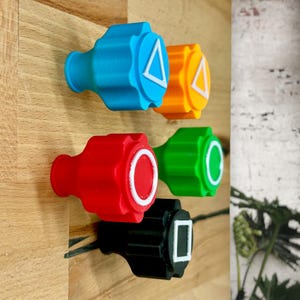 Op de afbeelding: Vier kleurrijke plastic knoppen met verschillende symbolen. De knoppen zijn blauw, oranje, rood en zwart. De symbolen zijn een driehoek, een cirkel, een vierkant en een cirkel met een streep erdoor.