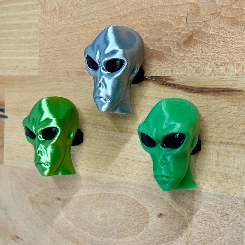 May include: Three alien head figurines, one silver, one green, and one lime green. The aliens have large black eyes and a serious expression.