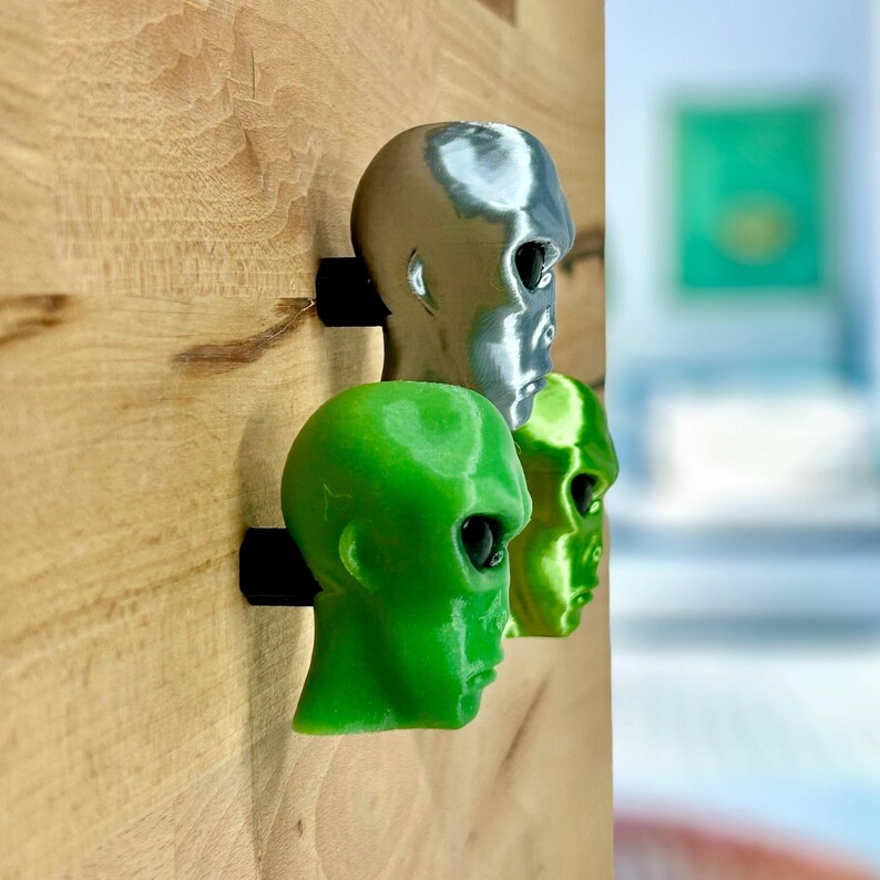 May include: Three alien head sculptures mounted on a wooden wall. The heads are made of 3D printed plastic and are painted in silver, green, and lime green.
