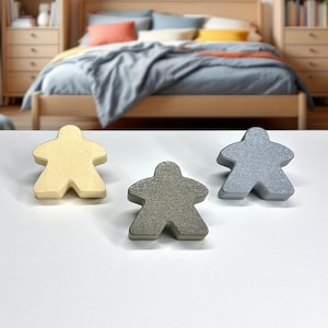 May include: Three wooden game pieces shaped like people. One is pale yellow, one is grey-green, and the other is silver. The pieces are on a white surface, with a bed in the background.