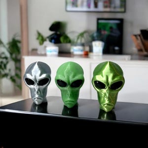 May include: Three alien head figurines, one silver, one green, and one lime green. The figurines are made of plastic and have a smooth, shiny finish.