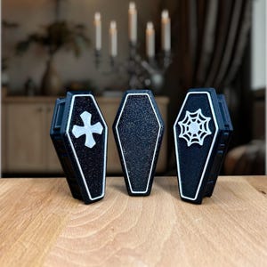 以下が含まれることがあります： 白いトリムが付いた、黒い棺の形をした3つの容器。1つは白い十字架、もう1つは空白の表面、3つ目は白いクモの巣のデザインです。容器は木製の表面にあります。