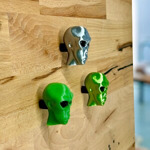 May include: Three alien head sculptures, one silver, one green, and one lime green, mounted on a wooden wall.