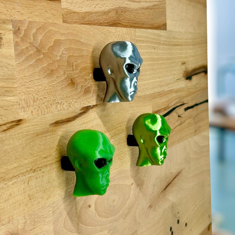 May include: Three alien head sculptures, one silver, one green, and one lime green, mounted on a wooden wall.