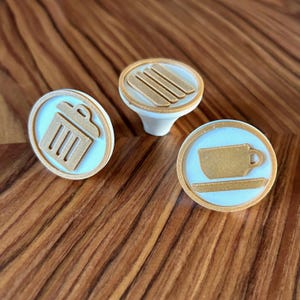 Op de afbeelding: Drie ronde keramische stempels in wit met een gouden rand. De stempels tonen pictogrammen van een prullenbak, een kopje en schoteltje, en een stapel van vier frietjes.