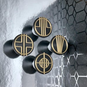 May include: Four black and gold round knobs with different geometric designs. The knobs are on a silver background.