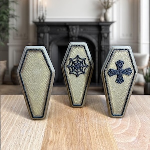 Pode incluir: Três pequenos ornamentos em forma de caixão de cor dourada com detalhes de glitter preto. Os ornamentos apresentam uma teia de aranha, uma cruz e um espaço em branco.