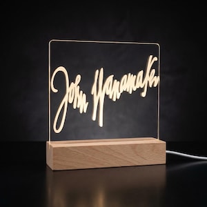 May include: A personalized LED night light with the name "John Watanabe" in a cursive font. The clear acrylic panel is illuminated by a warm light, set on a rectangular wooden base. The light is plugged in with a white cord.