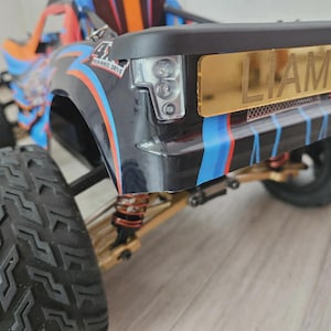 May include: A remote-controlled truck with a black, blue, and orange body featuring a gold-colored license plate on the rear bumper that displays the name LIAM in capital letters. The vehicle has large, knobby black tires and visible suspension.