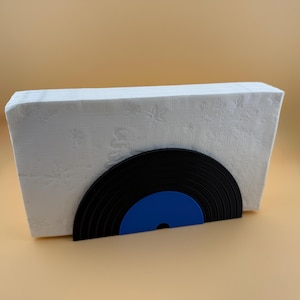 Op de afbeelding: Een witte, rechthoekige servethouder met een zwart en blauw vinylplaatontwerp. De houder is gemaakt van een stapel witte servetten en het plaatontwerp is zichtbaar aan de voorkant. De achtergrond is effen beige.