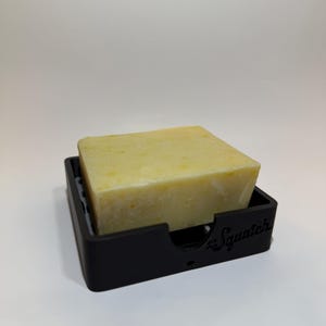 May include: A light green bar of soap in a black soap dish with the word "Squatch" printed on the dish.