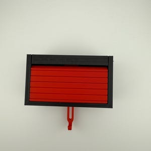 May include: A rectangular, black and red plastic object. The object has a black frame and a red, slatted front that appears to slide open. A red hook hangs from the bottom. The object is set against a white background.