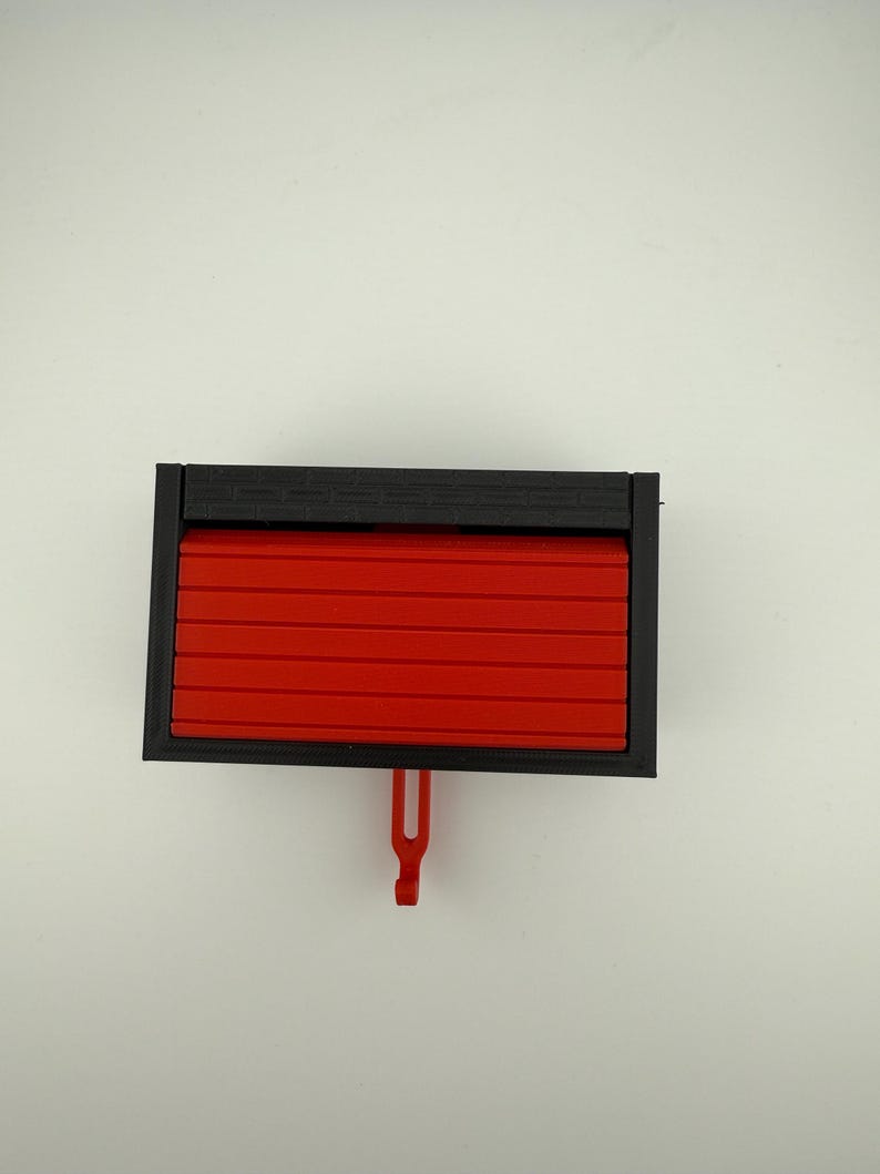 May include: A rectangular, black and red plastic object. The object has a black frame and a red, slatted front that appears to slide open. A red hook hangs from the bottom. The object is set against a white background.