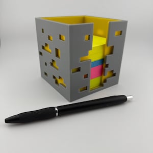 Op de afbeelding: Een grijze en gele vierkante bureau-organizer met een pixelontwerp, met een stapel kleurrijke plaknotities. Een zwarte pen ligt naast de organizer. De organizer is gemaakt van plastic en is ontworpen om plaknotities in te bewaren.