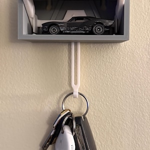 May include: A gray 3D-printed key holder shaped like a garage, with a small black toy car inside. A white hook hangs below, holding a keyring with keys and a small white device.