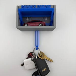 May include: A gray and blue key holder with the text "BRYAN'S GARAGE" above a miniature red car. A blue loop hangs below, holding keys, a key fob, and a small record-shaped keychain.