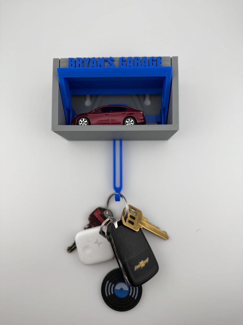 May include: A gray and blue key holder with the text "BRYAN'S GARAGE" above a miniature red car. A blue loop hangs below, holding keys, a key fob, and a small record-shaped keychain.