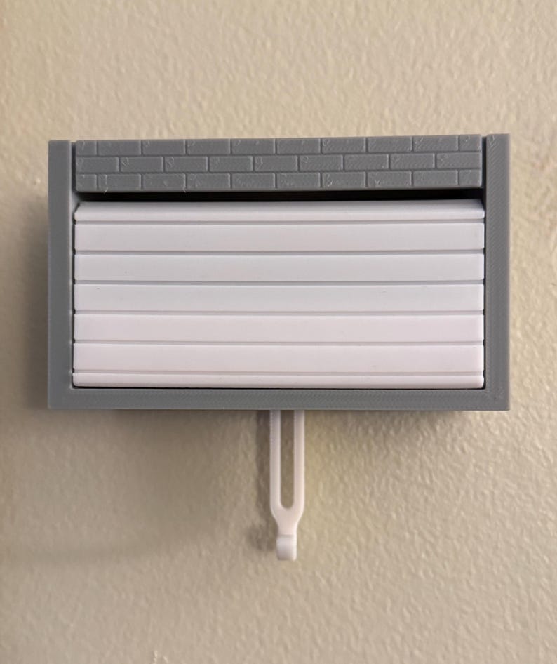 May include: A gray and white miniature garage door with a brick-like top detail and a white hook. The garage door is rectangular and has horizontal white slats. The hook is centered below the door.