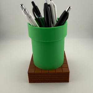 May include: A green pen holder in the shape of a video game pipe, set on a brown brick-effect base. The holder contains black and white pens. A fun and practical desk accessory.