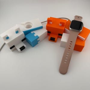 May include: A white, blue and orange 3D printed phone stand shaped like a fox, a dog and a cat. A rose gold Apple Watch is resting on the stand.