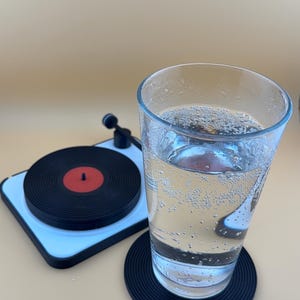 Puede incluir: Un vaso de agua con gas transparente sobre un posavasos con forma de disco negro y blanco. Un tocadiscos negro y blanco está en el fondo.