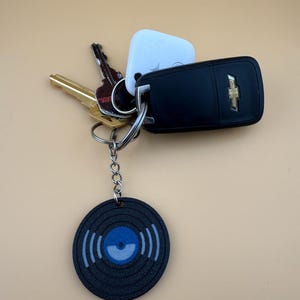 May include: A collection of keys and a black vinyl record keychain. The keychain features a black vinyl record design with blue and white accents. Other keys include a car key fob and a house key.