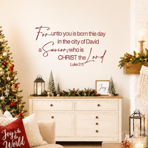 May include: A wall decal in dark red script lettering that reads: For unto you is born this day in the city of David a Savior, who is CHRIST the Lord Luke 2:11. The text is displayed on a plain wall above a white wooden cabinet in a room.