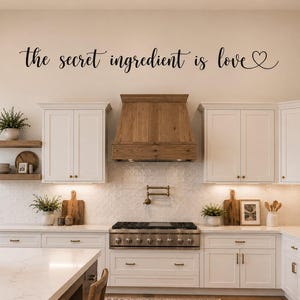 May include: A bright, modern kitchen with white cabinets, wooden accents, and a stainless steel range. The text "the secret ingredient is love" is written on the wall in black script. A black pendant light hangs from the ceiling.