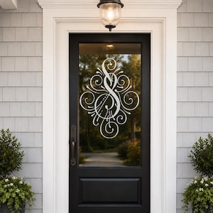 May include: A black front door with a glass panel featuring a white decorative monogram. The door is framed by white trim and set against gray siding. A black door handle and a hanging light fixture are visible.