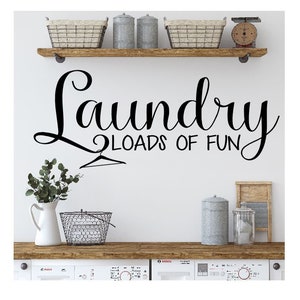 May include: A white wall with a black vinyl decal that reads "Laundry Loads of Fun" with a hanger graphic. The wall has a wooden shelf with three metal canisters and two wire baskets. There is a white countertop with a white pitcher, a wire basket, a metal canister, and a glass jar. Two white washing machines are visible below the countertop.