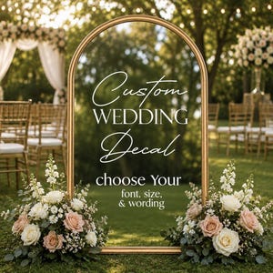 May include: A gold metal arch frame featuring white vinyl text that reads Custom WEDDING Decal choose Your font, size, & wording. The arch is decorated with two large floral arrangements of white and blush roses and greenery at the base.